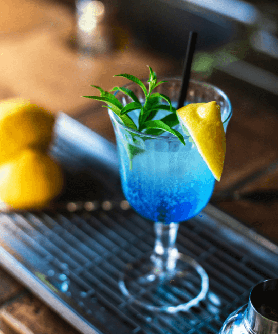 Copa de vidrio de bebida ready to drink XTASIS sabor Tropical Blue acompañado de un limón y hierbas de Xauxa